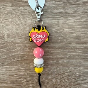 May include: A keychain with a heart-shaped charm and the words "slow burn" in pink and yellow. Below the charm are pink and white polka dot beads, a white bead, and a yellow bead. A black tassel hangs from the bottom.