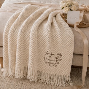 Può includere: Una coperta in maglia testurizzata color crema con frange sul bordo inferiore. La coperta è ricamata con i nomi Andre e Vivian, un piccolo cuore, un'illustrazione di una rosa e la data 20/7/2026 in filo marrone scuro.