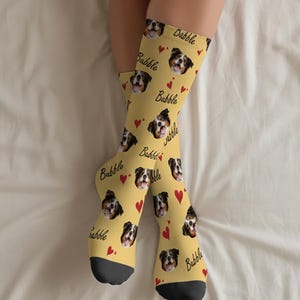 May include: Yellow socks featuring a repeating pattern of dog faces, red hearts, and the word "Dabble" in black script. The socks have black toes and heels.