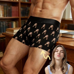 May include: Black boxer shorts featuring a repeating pattern of a woman's face. The shorts have a black waistband. The background includes a wooden desk and a bookshelf filled with books.