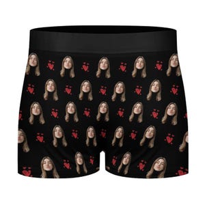May include: Black boxer briefs with a repeating pattern of a woman's face and red hearts. The boxer briefs have a black waistband and are made of a soft, comfortable material.