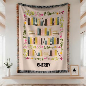 Può includere: Una coperta beige con un design a libreria con libri colorati e accenti floreali. La parola "CHERRY" è stampata in nero in basso. La coperta ha una frangia nera.