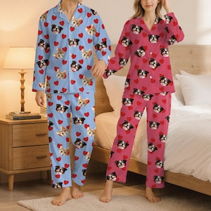 May include: Matching pajama sets in light blue and pink. The light blue set features a pattern of dog and cat faces with red hearts. The pink set has a similar design with dog faces and red hearts. Both sets include a button-down top and pants.