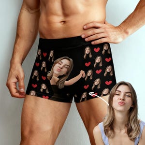 Könnte beinhalten: Schwarze Boxershorts mit einem sich wiederholenden Muster aus roten Herzen und Gesichtern. Auf der Vorderseite ist ein großes Bild eines Frauengesichts zu sehen. Die Boxershorts sind aus einem weichen, bequemen Material.