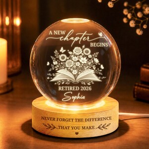 May include: A clear glass sphere engraved with the words "A New Chapter Begins" and "Retired 2026 Sophia". The sphere has a floral design and rests on a wooden base with the inscription "Never Forget The Difference That You Make."