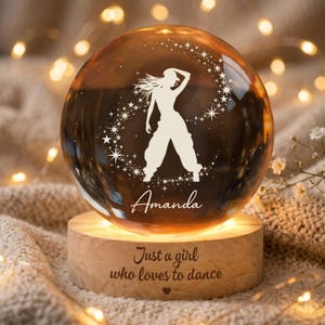 Può includere: Un globo di vetro trasparente con la silhouette di una ballerina e il nome "Amanda" inciso. Il globo poggia su una base di legno con la scritta "Just a girl who loves to dance". La base è illuminata da una calda luce.