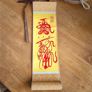 May include: A rolled-up, rectangular scroll with yellow and gold tones. The scroll features red Chinese calligraphy. A brown ribbon is attached to the top. The scroll is lying on a wooden surface.