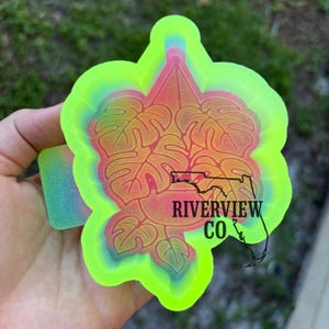 May include: A neon green and pink resin mold in the shape of a monstera leaf design. The mold has the text "RIVERVIEW CO" and a Florida outline. The mold is held in a hand.