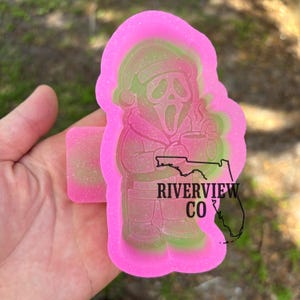Könnte beinhalten: Eine rosa-grüne Silikonform mit einer Cartoon-Figur von Ghostface mit Weihnachtsmütze, die eine Tasse hält. Die Form hat den Text "RIVERVIEW CO" und eine Florida-Umrisslinie.
