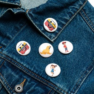 Może przedstawiać: Zestaw pięciu białych, okrągłych przypinek z kolorowymi ilustracjami z kreskówek. Projekty obejmują uśmiechniętą skrzynkę narzędziową, złotego retrievera, chłopca w czerwonej koszuli i dziewczynkę w niebieskiej sukience. Przypinki są umieszczone na niebieskiej dżinsowej kurtce.
