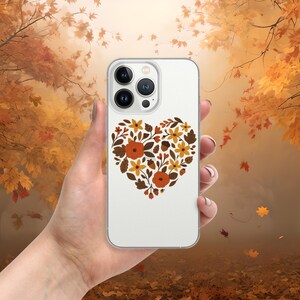 May include: A clear phone case featuring a heart-shaped floral design in shades of orange, brown, and yellow. The case is held against a blurred autumn background with falling leaves. The floral design includes flowers and leaves.