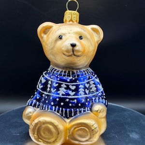 Puede incluir: Adorno navideño de cristal con forma de oso de peluche. El oso tiene la cabeza y las patas de color marrón dorado y lleva un jersey azul con diseños de copos de nieve blancos. El adorno tiene un lazo metálico dorado para colgarlo.