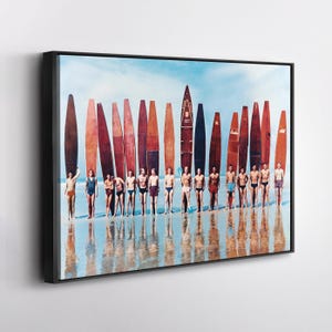 May include: A framed print featuring a vintage photograph of a group of people standing on a beach with surfboards. The surfboards are various shades of brown and red, and the people are wearing swimwear. The sky is blue, and the water reflects the scene.