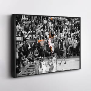 May include: Black and white framed print of a basketball game in action. A player in a red and white uniform is jumping to shoot the ball. The crowd is in the background.