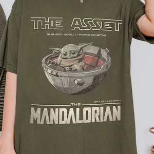 May include: Olive green t-shirt featuring a graphic of Grogu in a pod, with the text "THE ASSET" and "THE MANDALORIAN" in white. The shirt has a relaxed fit and short sleeves.