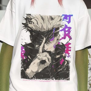 May include: White t-shirt with a graphic of an anime character in black, white, and purple. The shirt has black trim on the sleeves. The person in the image is wearing a purple beanie and has pink hair.