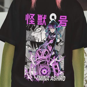 Könnte beinhalten: Schwarzes T-Shirt mit einem Manga-Grafikdesign, japanischem Text und dem Namen "MINA ASHIRO". Das Design zeigt eine weibliche Figur in einem futuristischen Outfit mit lila und weißen Akzenten. Die Person trägt eine beige Mütze und hat rosa Haare.