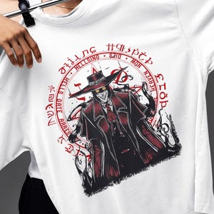 Puede incluir: Camiseta blanca con un gráfico rojo y negro de un personaje con abrigo largo y sombrero. El diseño incluye texto en un patrón circular alrededor de la imagen. La camiseta está sujeta por una persona.