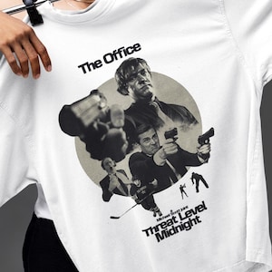 May include: White t-shirt featuring a graphic from "The Office" with the text "Threat Level Midnight." The design includes images of characters in action poses. The shirt is held up by a person.