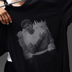 May include: Black t-shirt with a grayscale image of a person. The person is wearing a white t-shirt. The t-shirt is being held up by a person wearing a black and white patterned bucket hat.