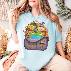 May include: Light blue t-shirt featuring a graphic of a purple backpack filled with Toy Story characters. The backpack has the text "TOY STORY 5" on it. The shirt is a short-sleeved crew neck.