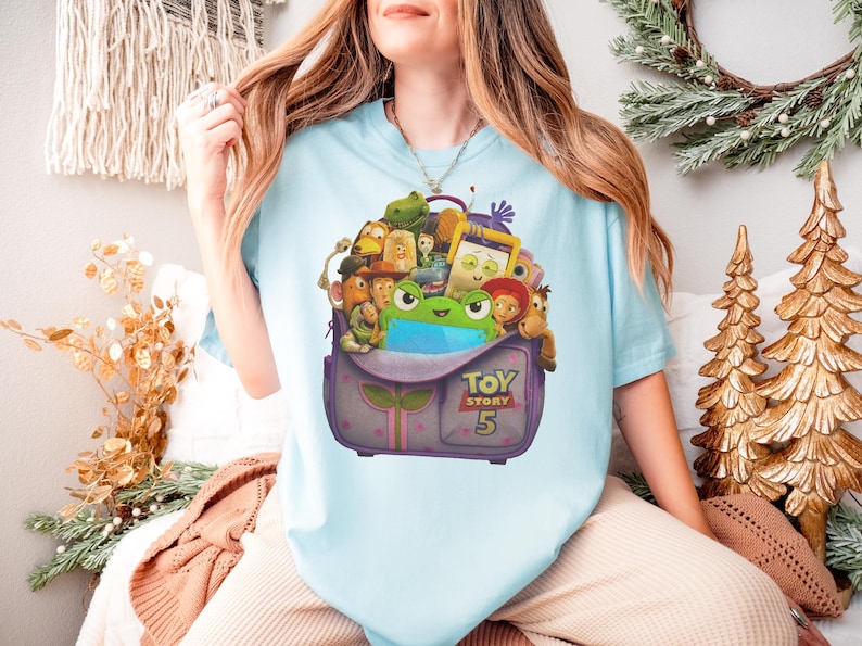 May include: Light blue t-shirt featuring a graphic of a purple backpack filled with Toy Story characters. The backpack has the text "TOY STORY 5" on it. The shirt is a short-sleeved crew neck.