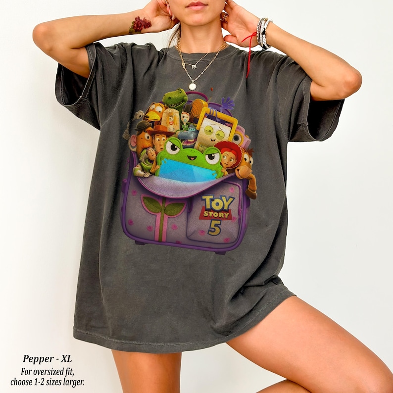 May include: Dark gray oversized t-shirt featuring a purple backpack graphic filled with Toy Story characters. The backpack has the text "TOY STORY 5". The shirt is a relaxed fit and made of a soft material.