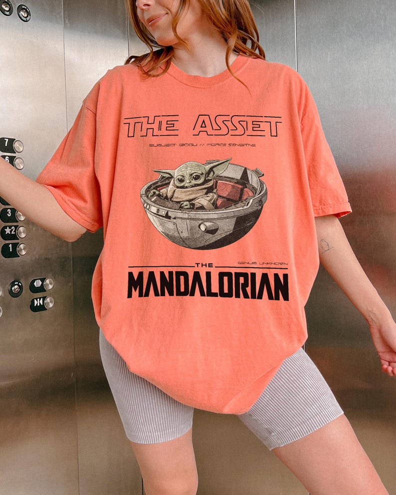 May include: Coral-colored t-shirt featuring a graphic of Grogu in a pod from The Mandalorian. The text "THE ASSET" and "THE MANDALORIAN" are printed above and below the image in black. The shirt has short sleeves and a relaxed fit.