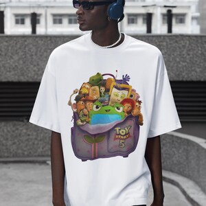 May include: White t-shirt featuring a colorful graphic of characters from the animated film "Toy Story 5". The graphic includes a frog, various toys, and the film's title. The shirt is worn by a person wearing sunglasses and headphones.