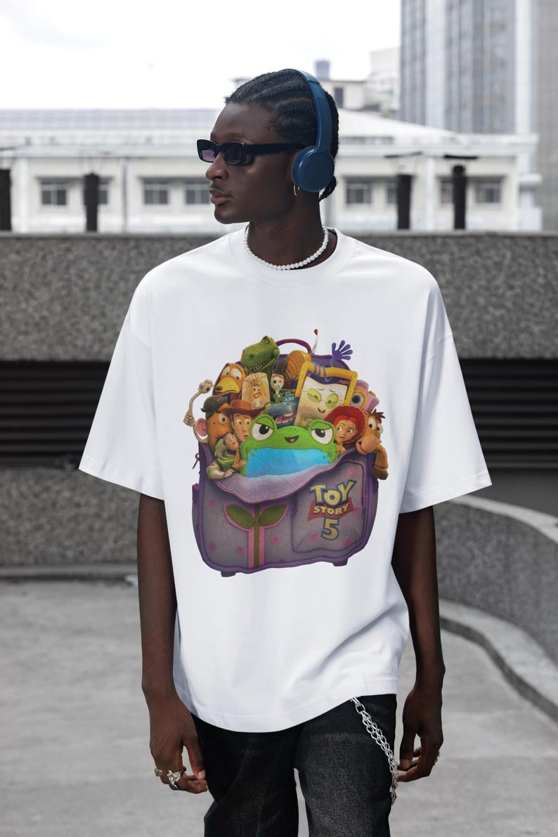 May include: White t-shirt featuring a colorful graphic of characters from the animated film "Toy Story 5". The graphic includes a frog, various toys, and the film's title. The shirt is worn by a person wearing sunglasses and headphones.