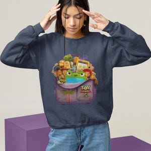 May include: Navy blue sweatshirt featuring a colorful graphic of characters from the movie Toy Story 5. The graphic is inside a purple backpack with the text "Toy Story 5". The sweatshirt is a crew neck style.