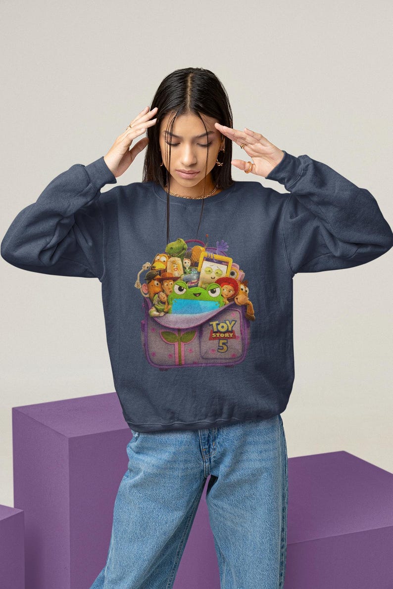 May include: Navy blue sweatshirt featuring a colorful graphic of characters from the movie Toy Story 5. The graphic is inside a purple backpack with the text "Toy Story 5". The sweatshirt is a crew neck style.