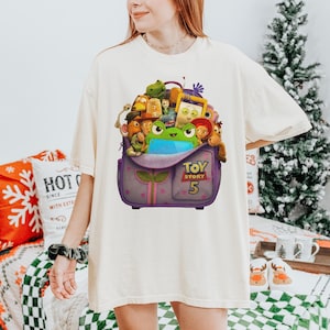 May include: Cream-colored t-shirt featuring a graphic of a purple backpack overflowing with Toy Story characters. The backpack has the text "TOY STORY 5" on the front. The shirt is a relaxed fit.