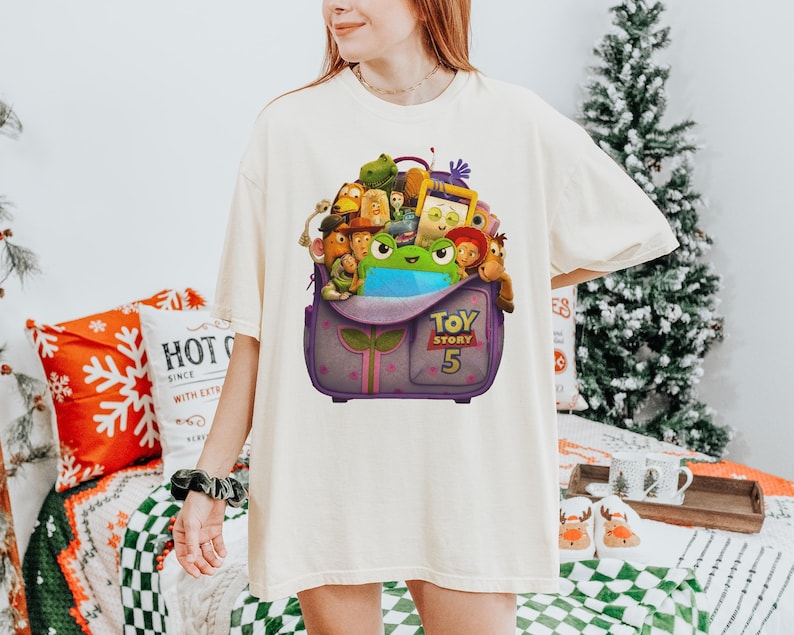 May include: Cream-colored t-shirt featuring a graphic of a purple backpack overflowing with Toy Story characters. The backpack has the text "TOY STORY 5" on the front. The shirt is a relaxed fit.