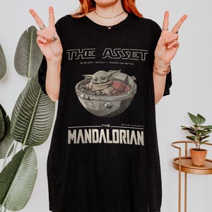 May include: Black t-shirt featuring a graphic of Grogu in a pod from The Mandalorian. The text "THE ASSET" is above the image, with "SUBJECT GROGU / FORCE SENSITIVE" below it. The text "THE MANDALORIAN" is below the image.