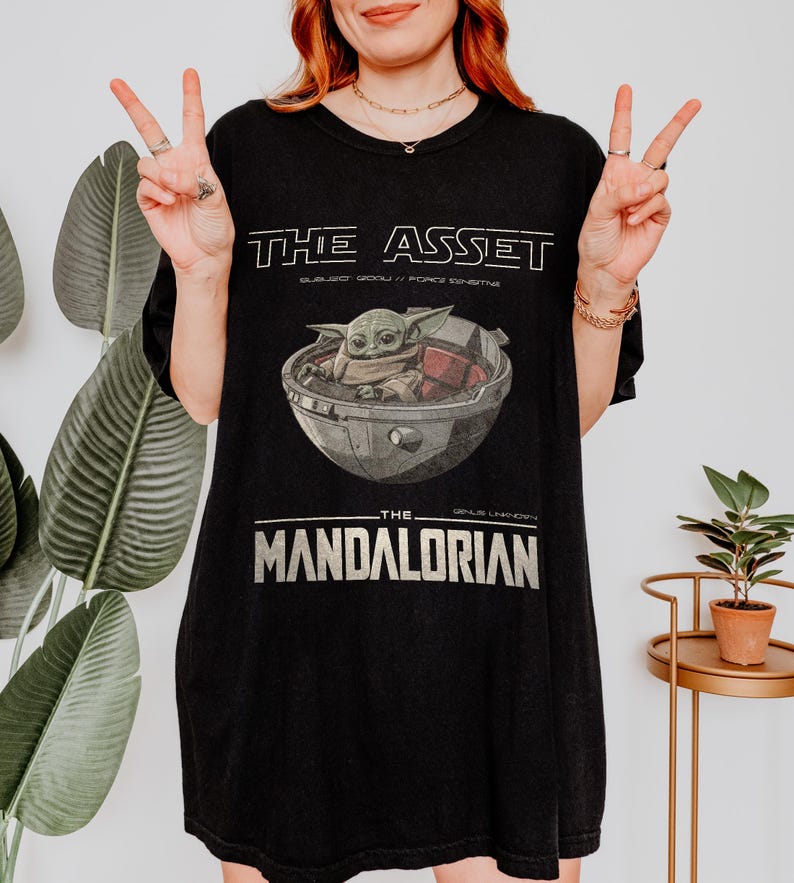 May include: Black t-shirt featuring a graphic of Grogu in a pod from The Mandalorian. The text "THE ASSET" is above the image, with "SUBJECT GROGU / FORCE SENSITIVE" below it. The text "THE MANDALORIAN" is below the image.