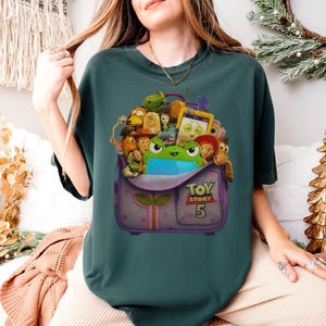 May include: Teal-colored t-shirt featuring a graphic of a purple backpack overflowing with animated characters from the Toy Story franchise. The backpack has the text "TOY STORY 5" on it. The shirt is a comfortable, casual style.