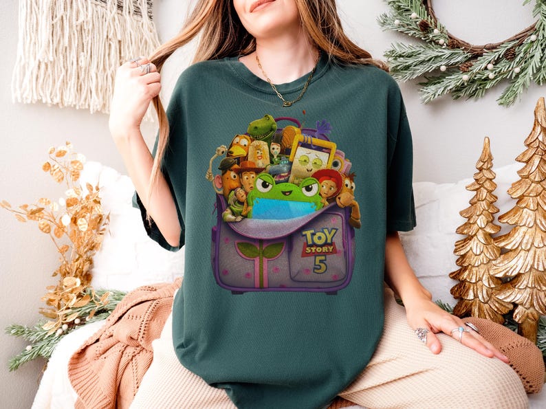 May include: Teal-colored t-shirt featuring a graphic of a purple backpack overflowing with animated characters from the Toy Story franchise. The backpack has the text "TOY STORY 5" on it. The shirt is a comfortable, casual style.