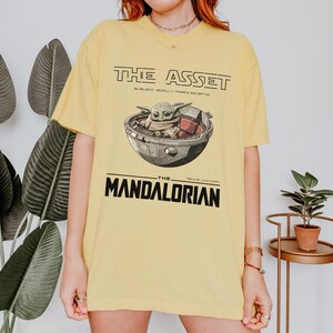 May include: Yellow t-shirt featuring a graphic of Grogu in a pod, with the text "THE ASSET" above and "THE MANDALORIAN" below. The design is in black and white, with the text in a bold, sans-serif font.
