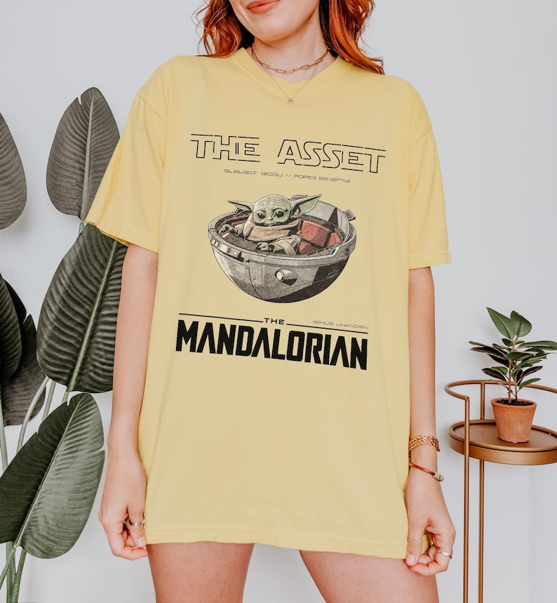 May include: Yellow t-shirt featuring a graphic of Grogu in a pod, with the text "THE ASSET" above and "THE MANDALORIAN" below. The design is in black and white, with the text in a bold, sans-serif font.