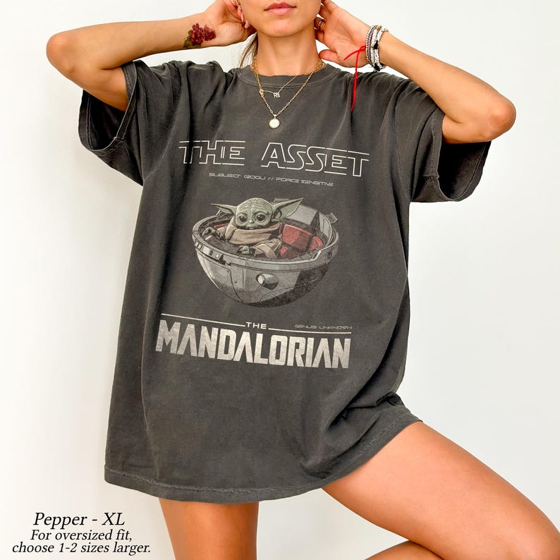 May include: Dark grey t-shirt featuring a graphic of Grogu in a pod, with the text "THE ASSET" and "THE MANDALORIAN" in white. The shirt is oversized and has short sleeves. The text "Pepper - XL" is at the bottom.