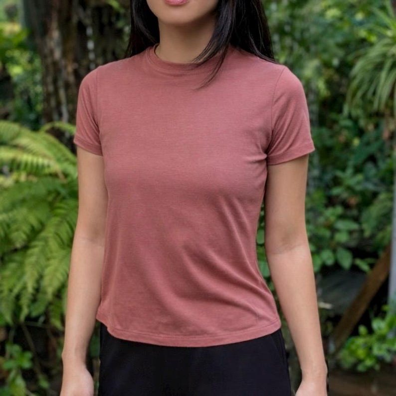 May include: A solid dusty rose short-sleeved t-shirt with a crew neckline. The garment features a simple, plain design with a fitted silhouette and a smooth, matte fabric texture, worn with plain black bottoms that are visible at the waistline.