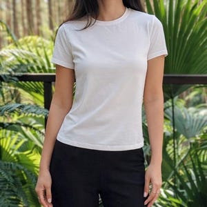 May include: A plain white short-sleeved t-shirt with a crew neckline, paired with solid black trousers. The t-shirt features a simple, fitted silhouette with clean lines and a smooth fabric texture, providing a neutral and versatile appearance.