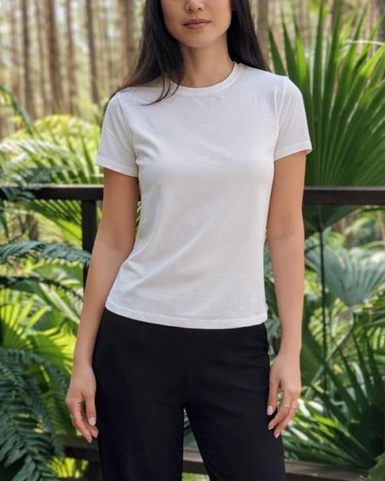 May include: A plain white short-sleeved t-shirt with a crew neckline, paired with solid black trousers. The t-shirt features a simple, fitted silhouette with clean lines and a smooth fabric texture, providing a neutral and versatile appearance.