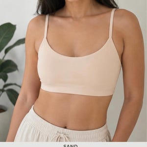 May include: A sand-colored bralette with thin shoulder straps and a scoop neckline. The text below the garment reads: SAND, Bralette 2.0 - More coverage, True to size. The bralette features a smooth, solid-colored fabric design.