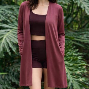 May include: A burgundy three-piece activewear set. The set includes a long-sleeved cardigan, a crop top, and shorts. The cardigan has pockets and a draped front. The crop top has a scoop neck. The shorts are high-waisted.