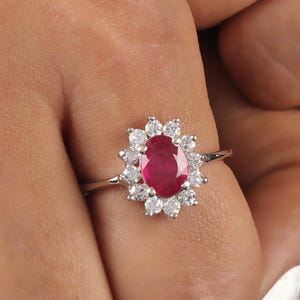 May include: A silver ring featuring a large oval ruby gemstone surrounded by a halo of smaller, clear gemstones. The ring is set on a delicate band, creating a classic and elegant design. The ring is shown on a person's finger.