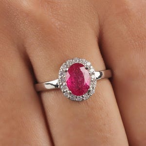 May include: A silver ring featuring an oval-cut ruby gemstone surrounded by a halo of small, round diamonds. The ruby is a vibrant red color, and the diamonds sparkle against the silver band. The ring is a classic and elegant piece of jewelry.