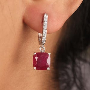 May include: A silver hoop earring with a square, deep red gemstone. The hoop is encrusted with small, clear stones. The gemstone is a vibrant, saturated red color. The earring is shown on a person's ear.