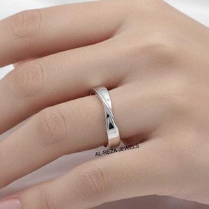 Puede incluir: Un anillo plateado con un diseño retorcido se muestra en una mano. El anillo tiene un acabado pulido y una forma suave y curvada. El fondo es una tela blanca y sedosa. El anillo es una joya.